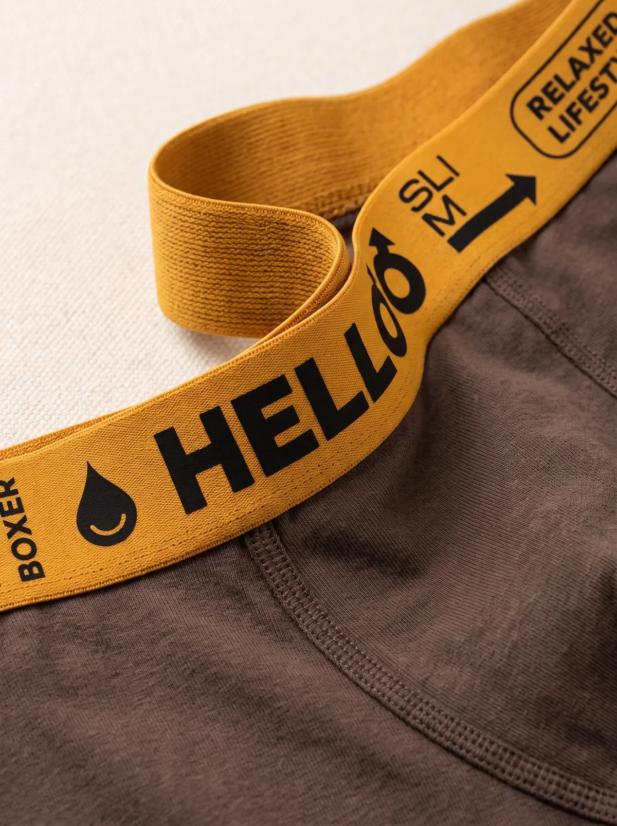 A mustard-yellow woven waistband lifted away from a brown cotton boxer brief, showing the soft elastic